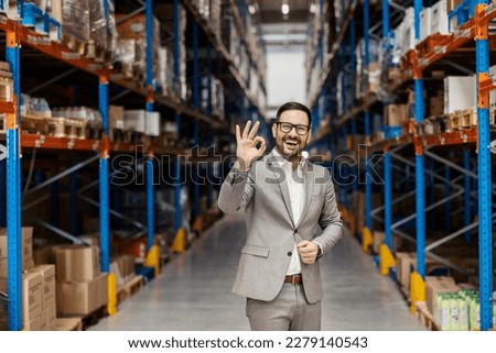An executive is showing okay gesture while smiling at the camera in export and shipping firm in warehouse.
