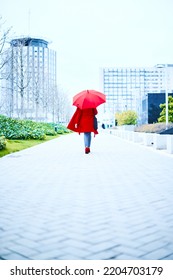 Executive Woman Walking Under A Red Umbrella In A Rainy Day, She Is Dressed In Blue Jeans And Red Coat Looking To The Side. Background Of An Office Building. Copy Space Business Executive Woman.