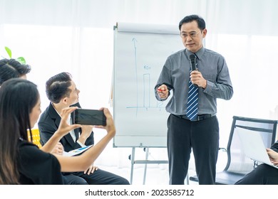 Executives hold a microphone presenting work plans in the conference room and a female employee uses a smartphone to take pictures in the conference room.