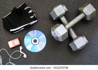 Exercise Equipment On Black Mat. Gloves, Music, DVD And Dumbbells.