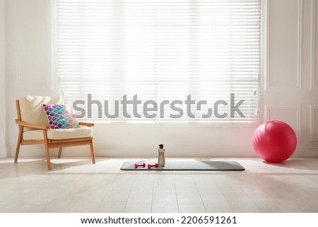 Exercise mat, dumbbells, bottle and fitness ball near armchair in spacious room