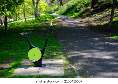 An Exercise Spin Bike Placed In A Public Park. Manual Exercise Bike For Passers By To Cycle On. Next To The Footpath And Green Grass. Sunny Spring Day. No People. Concept For Fitness And Wellbeing.