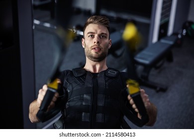Exercising arms and chest at EMS training. A young attractive man in a suit for electrical muscle stimulation doing exercises on hanging straps in the gym, TRX. Strength, strong movement