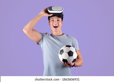 Exercising With Virtual Reality Concept. Young Athletic Guy In VR Headset Holding Soccer Ball On Lilac Studio Background. Positive Young Sportsman Using Modern Technologies For Football Workout