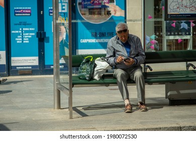 Exeter, England, UK -  04.23.2021: An Elderly Caucasian Man Sits At A Bus Stop, While On His Phone.