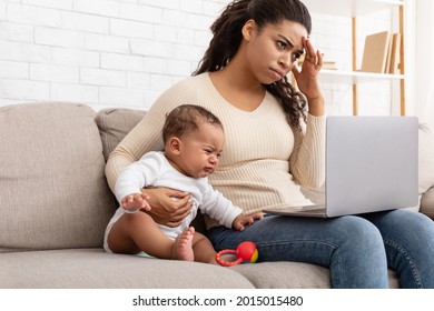 Exhausted African American Mother Hugging Crying Baby Using Laptop Browsing Internet Sitting At Home. Mom Trying To Work Online On Computer While Infant Son Screaming Feeling Bad