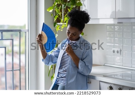 Exhausted African American woman standing in kitchen holding paper fan and feeling unwell due to stuffiness. Young black girl breathing uses hand fan to soothe herself after heatstroke from summer sun