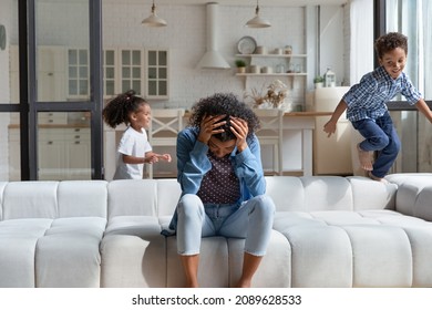 Exhausted African Mother Sit On Sofa Holding Head Suffers From Headache Due To Noisy Active Misbehaved Little Children Running Around At Home. Upbringing Difficulties, Divorced Depressed Mom Concept