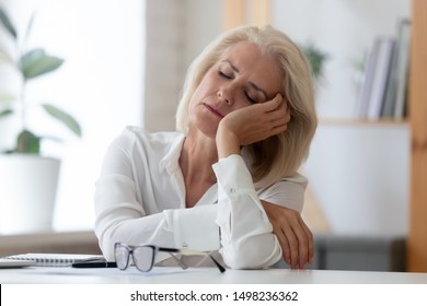 Exhausted aged woman worker sit at office desk fall asleep distracted from work, tired senior businesswoman feel fatigue sleeping at workplace taking break dreaming or visualizing