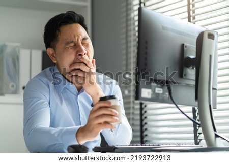 Exhausted Asain business man using laptop computer working in office. Young employee male worker sitting on table feel sleepy and yawning tired from overwork job and drinking hot coffee in workplace. Foto stock © 