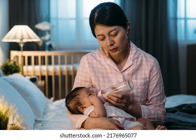 Exhausted Asian Mother Is Sleeping Dozing Off While Sitting At Bedside Giving Her Young Baby Bottled Milk Late At Night In A Dark Bedroom With Dim Light At Home.