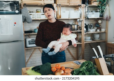 Exhausted Asian New Mom Is Looking Away With A Frown While Having Trouble Dealing With Her Angry Wailing Baby She’s Holding In The Kitchen At Home.