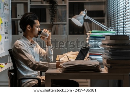 Exhausted Asian young businessman overworking late at night in office. Attractive busy male employee worker feel tired of many paper work on table and use computer plan project at corporate workplace.