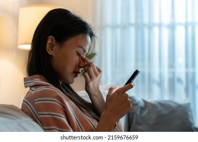 Exhausted Asian Young Woman Hurt Eye While Using Mobile Phone At Night. Attractive Girl Sitting On Sofa Feeling Visual Fatigue And Eye Strain Tired From Overwork And Massage Dry Irritable Eye At Home.