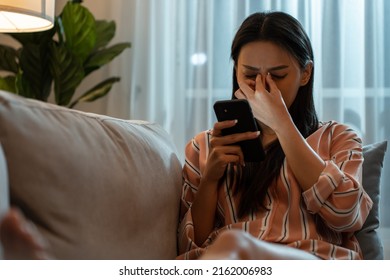 Exhausted Asian Young Woman Hurt Eye While Using Mobile Phone At Night. Attractive Girl Sitting On Sofa Feeling Visual Fatigue And Eye Strain Tired From Overwork And Massage Dry Irritable Eye At Home.