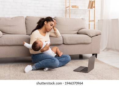 Exhausted Black Mother Holding Crying Baby And Touching Head Suffering From Headache Sitting At Laptop On Floor At Home. Motherhood Burnout Concept. Tired Mother Posing With Shouting Child