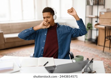 Exhausted black teenage boy in headphones yawning, showing signs of tiredness and need for rest during study session, with laptop and notes in front