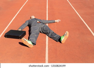 An Exhausted Business Man In Gray Suit With Green Shirt And Tie, Black Briefcase And Broken Green Running Shoes Lying On A Running Track.