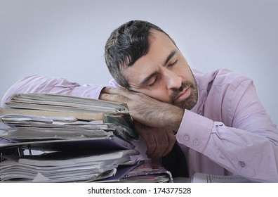 Exhausted businessman with a beard asleep at his desk with his head resting on a large stack of office files and paperwork