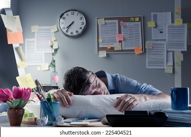 Exhausted businessman sleeping at workplace with a pillow on his desk. - Shutterstock ID 225404521