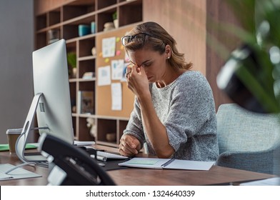 Exhausted Businesswoman Having A Headache At Office. Mature Creative Woman Working At Office Desk Feeling Tired. Stressed Casual Business Woman Feeling Eye Pain While Overworking On Desktop Computer.