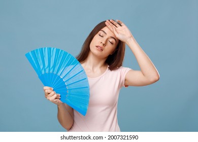 Exhausted Caucasian Young Woman With Fan Touching Her Forehead, Feeling Bad Because Of Summer Heat, Suffering From High Temperature Outside, Blue Studio Background, Copy Space.