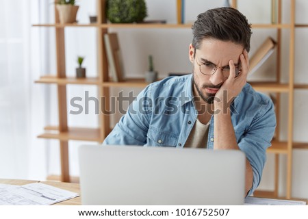 Exhausted embarrassed male reads some information on laptop computer, being tired and overworked after hard work at office all day long, doesn`t understand how make project, being frustrated