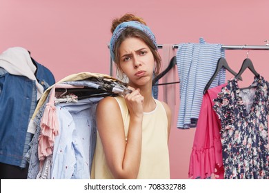 Exhausted female model blowing cheeks and having tired expression while standing in fitting room, holding hangers with clothes on shoulder, frowning face with dissatisfaction. Tiredness and shopping