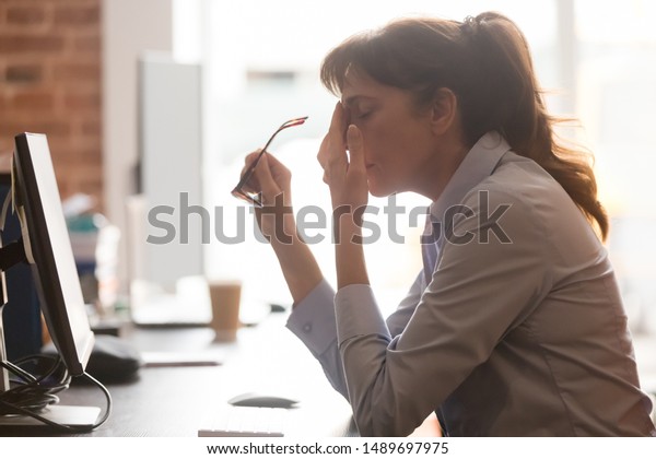 Exhausted female worker sit at office desk take\
off glasses feel unwell having dizziness or blurry vision, tired\
woman employee suffer from migraine or headache unable to work.\
Health problem\
concept