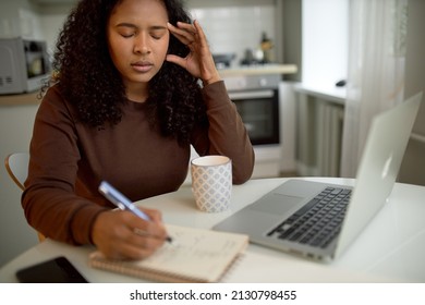 Exhausted Frustrated Female Of Dark Ethnicity Holding Temples Feeling Pain Or Having Headache,working From Home, Online, Writing Down Information In Her Copy Book, Drinking Hot Tea