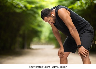 Exhausted Handsome African American Young Guy Jogging In Public Park, Having Break While Getting Ready For Marathon, Listening To Music, Using Earbuds, Panorama With Copy Space