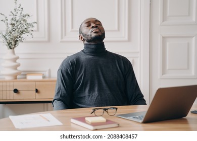 Exhausted Home Office Employee Afro American Male Sitting In Front Of Laptop Taking Off Glasses Closing Eyes And Throwing Head Back. Tired Young Man Trying To Recover To Continue Remote Work Online