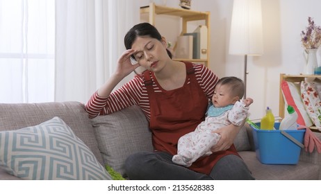exhausted housewife is coaxing her baby girl by patting while suffering headache after doing tiring housework on the couch in the living room.