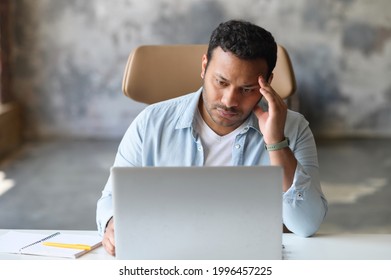 Exhausted indian office employee sits at the desk with a laptop and holds head with hand, tired guy feels burnout and depression, does not have time to complete the project before the deadline