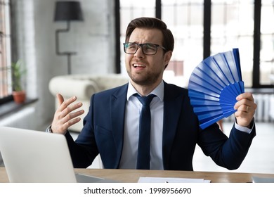 Exhausted Male Office Employee Suffering From Heat, Hot Stuffy Air. Business Man Cooling With Handheld Fun While Working On Summer Day In Office Without Conditioner. Conditioning, Ventilation Concept