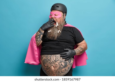 Exhausted male superhero yawns and covers mouth stands with big tattooed belly indoor being tired of doing different things models against blue background. Plump man dresses for costume party