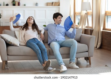 Exhausted Man And Woman Waving Blue Paper Fans, Breathing, Resting Sitting On Couch In Living Room, Overheated Tired Couple Suffering From Hot Summer Weather, Heating At Home, Feeling Discomfort