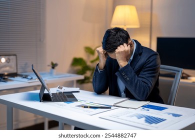 Exhausted manager having a headache while analyzing marketing reports and financial charts on laptop, working until late at night in office