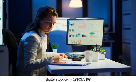 Exhausted manager trying to finish business project respecting deadline working at night in front of computer taking notes writing on notebook. Tired employee overtime sitting at desk in workplace