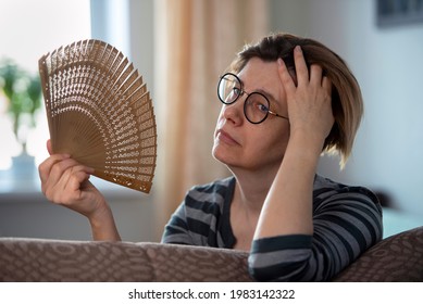 Exhausted middle aged woman waving her fan, suffering from menopausal symptoms, experiencing hot flush.  - Shutterstock ID 1983142322
