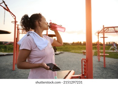 Exhausted middle-aged sportswoman in pink t-shirt with white terry towel drinks water, holds a smartphone and rests after an evening workout at sunset on the sports ground - Powered by Shutterstock