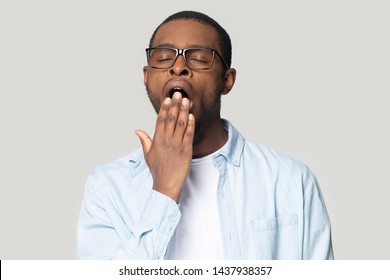 Exhausted millennial african American man in glasses isolated on grey studio background sigh having sleep deprivation, tired black male in spectacles yawn cover mouth feel sleepy, lack rest or relax