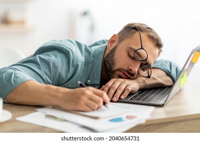 Exhausted millennial man sleeping on his office desk, next to laptop and documents, tired of overworking. Young male workaholic suffering from chronic fatigue at workplace