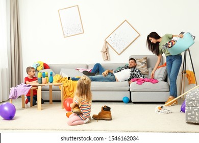 Exhausted Mother Trying To Clean Up Mess Made By Children And Lazy Father In Room