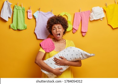 Exhausted mother yawns and wants to sleep, poses with newborn baby wrapped in blanket, tired of doing domestic chores, has much work as cares about infant. Parenthood, tiredness and moterhood