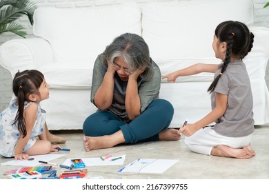 Exhausted Old Elderly Grandmother Sit On Floor In Living Room And Feel Unwell Tired From Little Children Running And Playing Loud, Suffer From Headache, Female Nanny Feels Exhausted By Noisy Kids