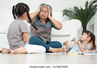 Exhausted Old Elderly Grandmother Sit On Floor In Living Room And Feel Unwell Tired From Little Children Running And Playing Loud, Suffer From Headache, Female Nanny Feels Exhausted By Noisy Kids