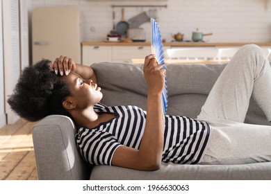 Exhausted Overheated Black Female Lying On Sofa With Paper Fan Suffer From Hot Temperature Inside And Headache. Shocked African Woman Breathe Fresh Cooled Air To Calm Down Panic Attack Alone At Home