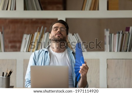 Exhausted overheated businessman in glasses waving paper fan, sitting at desk in office, young man suffering from overheating, high temperature, hot summer weather without conditioner at workplace