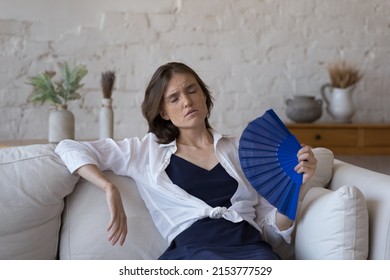 Exhausted Overheated Girl Feeling Hot At Home Due To Summer Weather, Too High Air Temperature In Apartment, Feeling Discomfort, Heat Stroke, Resting On Sofa, Waving Paper Handheld Fan Blowing Cool Air